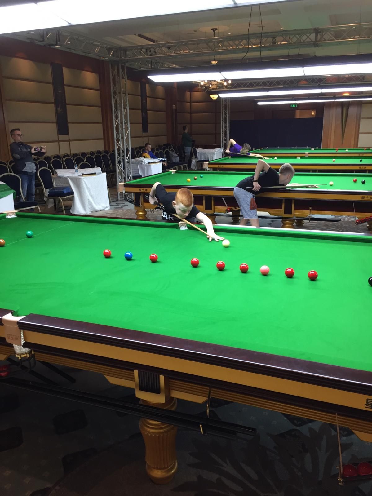 Snookerwedstrijd aan de gang. Spelers buigen zich over de tafel en leggen hun slagen klaar. Groene tafel met rode, blauwe en witte ballen.