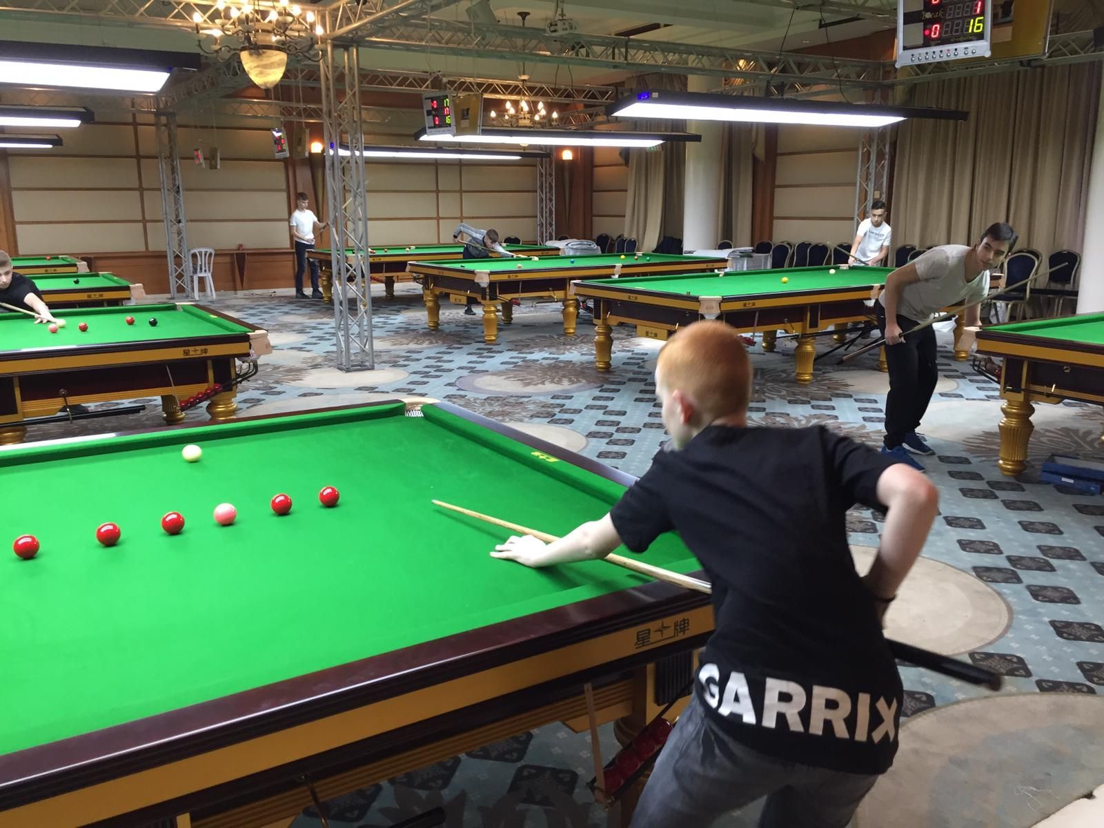 Een jonge jongen met rood haar bereidt zich voor op een snookerbal in een felverlichte snookerhal, met in het zicht verschillende andere tafels.