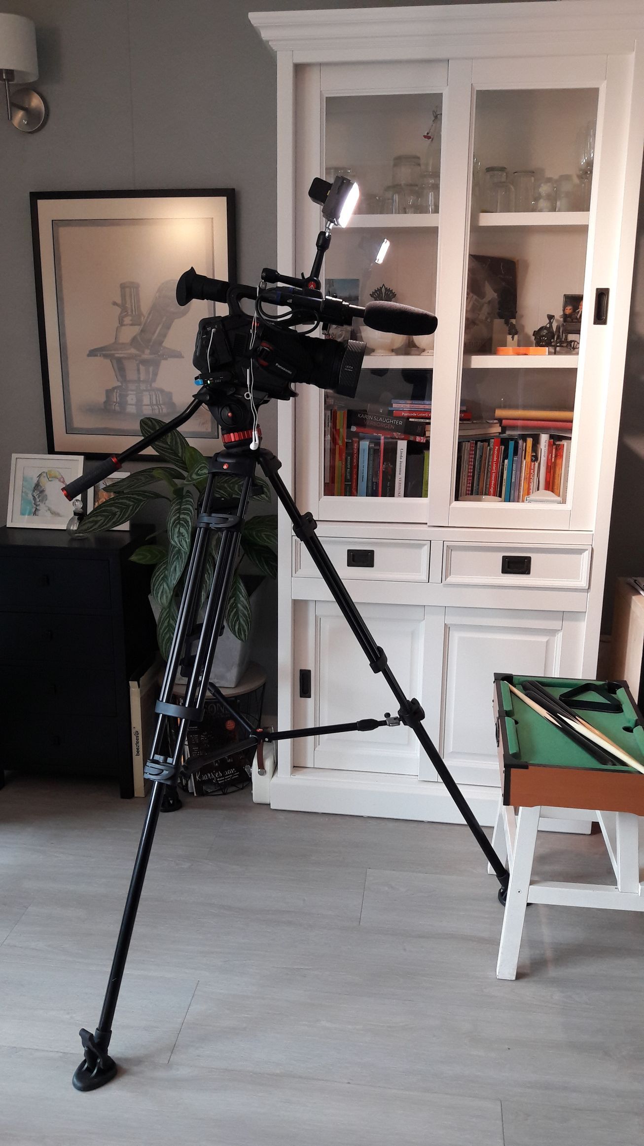 Een camera op een statief staat voor een witte kast met boeken en decoraties erin.