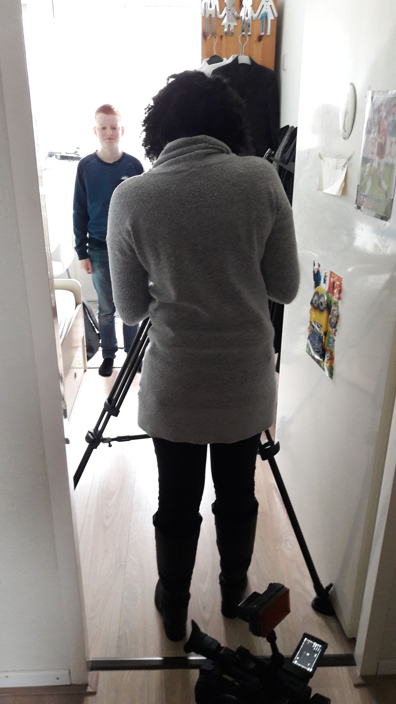 Vrouw filmt een man in een deuropening. Camera op statief. Man lacht. Lichte kamer.