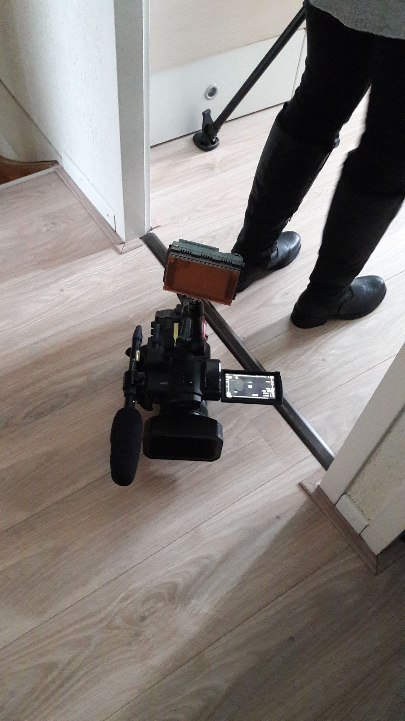 Een camera op een statief staat in een deuropening. Ernaast zijn de benen van een persoon zichtbaar die op de lichte houten vloer staat.