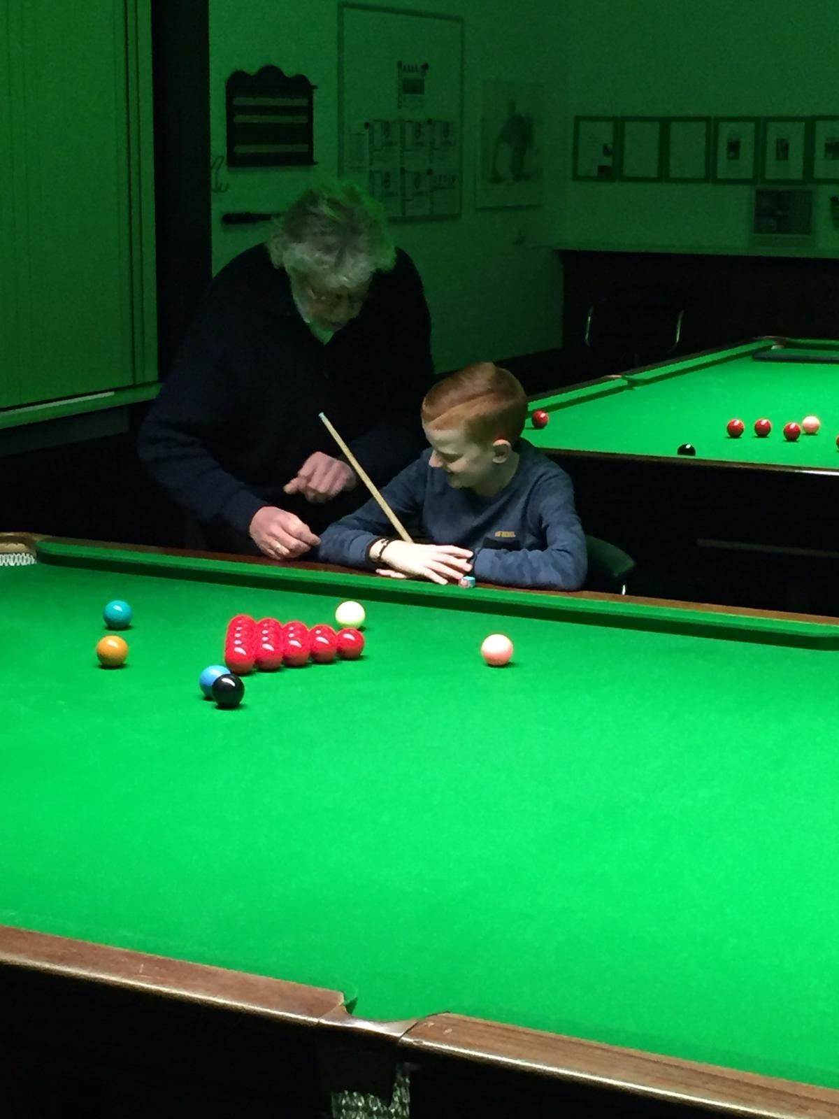 Een jongen krijgt snookerles van een man aan een groene tafel in een schemerige kamer.