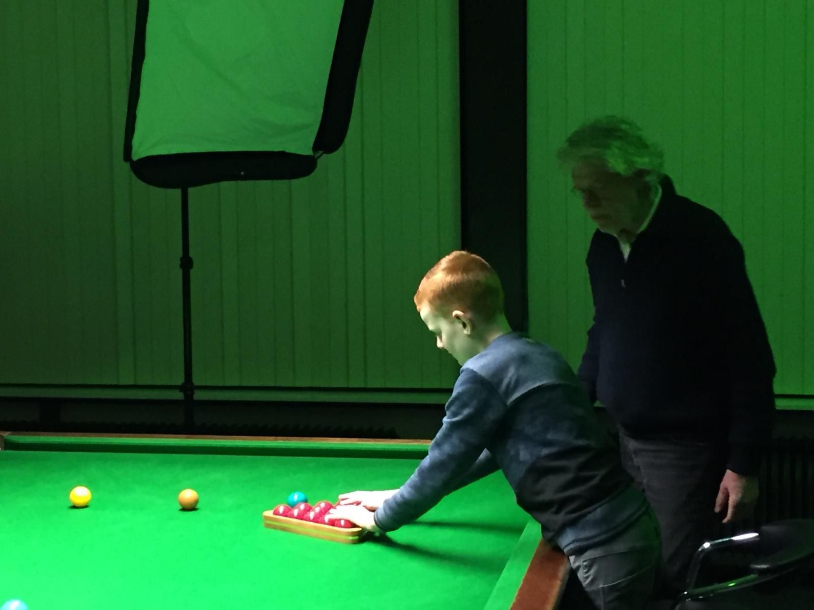 Een jonge jongen in een blauw shirt oefent snooker met een oudere man in een groen verlichte kamer.