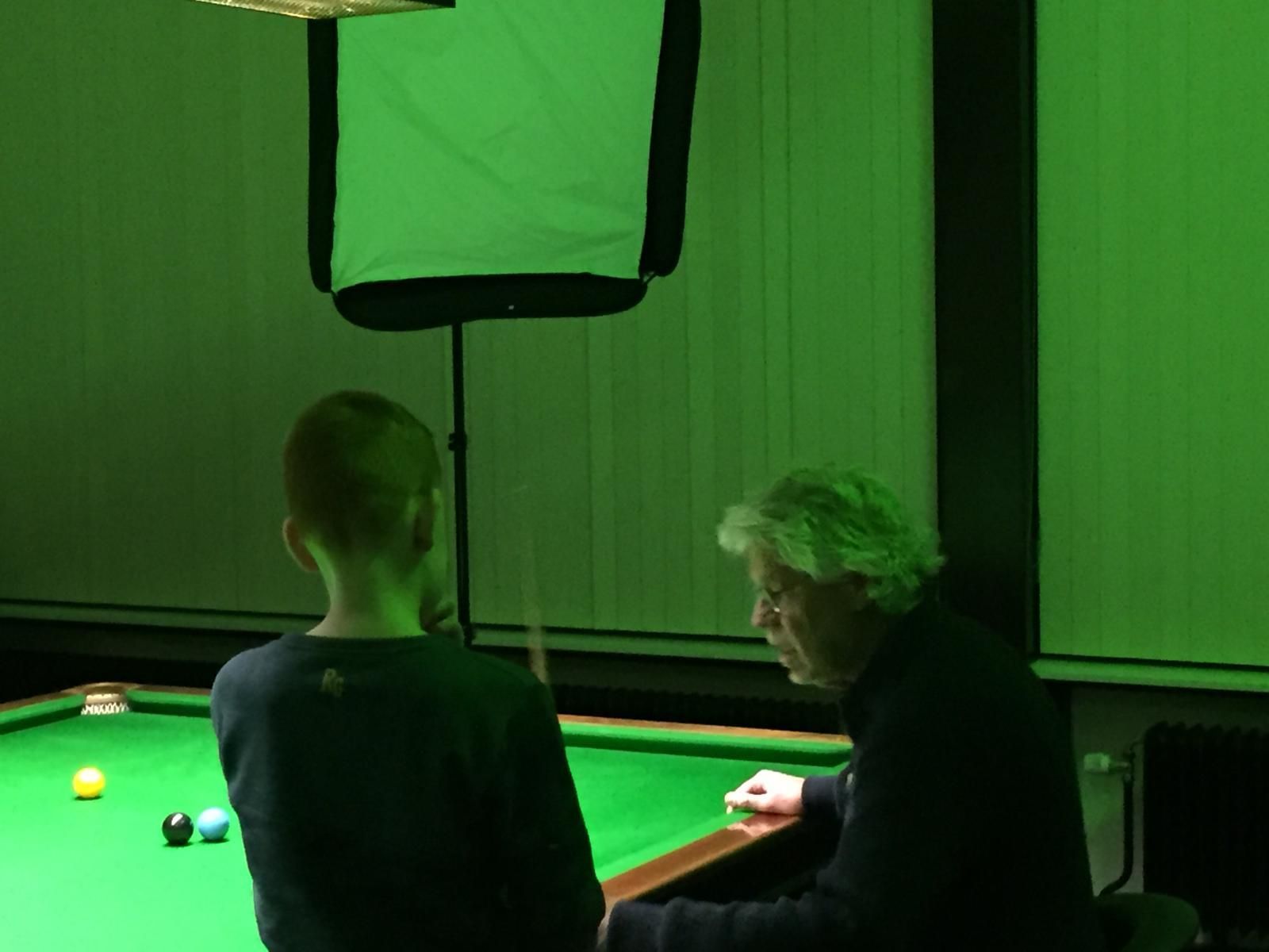 Een man coacht een kind op een groene snookertafel, onder groen licht.