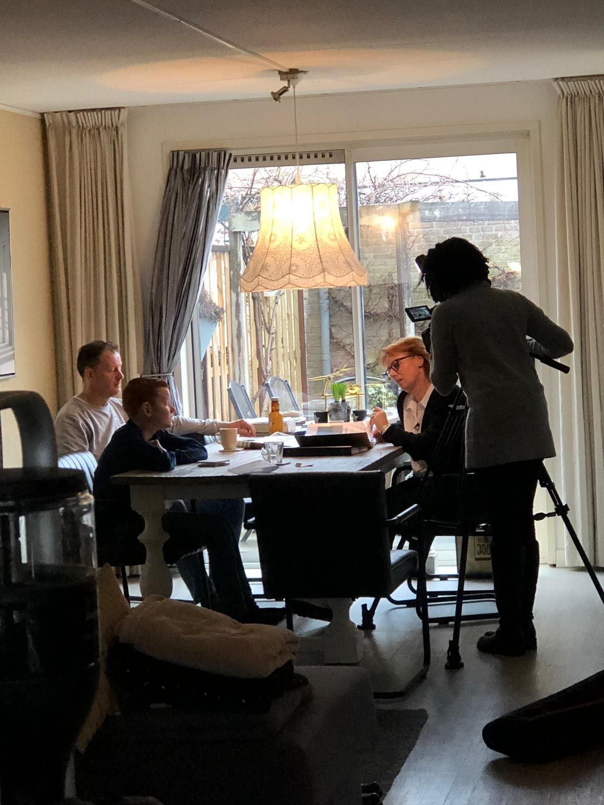 Mensen filmen een scène aan een tafel in een huis, met een cameraman, een lamp en uitzicht van buitenaf.