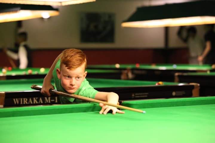 Een jonge roodharige bereidt zich voor op een stoot in een biljartzaal, met gefocuste ogen, en houdt een keu vast boven een groene tafel van vilt.