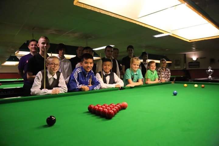Groep kinderen en tieners aan een snookertafel, lachend, vooruitkijkend. Rode en andere gekleurde ballen. Groene tafel.