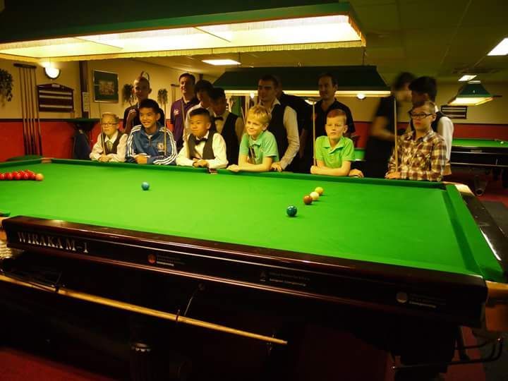 Kinderen en volwassenen verzamelen zich rond een groene snookertafel en kijken in clubverband naar de wedstrijd.