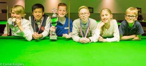 Zes kinderen poseren op een groene pooltafel, sommigen dragen medailles en houden een trofee vast, lachend.