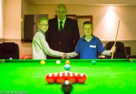 Twee jongens schudden elkaar de hand boven een snookertafel, terwijl een volwassene toekijkt. Er liggen snookerballen klaar.