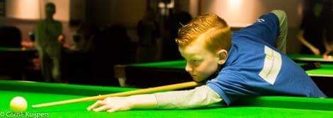 Jonge jongen met rood haar speelt snooker, geconcentreerde uitdrukking, groene tafel, keu, witte bal.