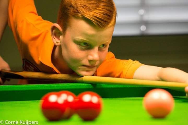 Een jonge jongen met rood haar richt geconcentreerd met een keu op rode en roze ballen op een groene tafel.