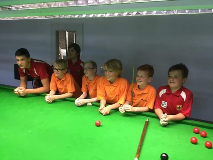 Kinderen in oranje en rode shirts kijken achter een groene tafel naar een snookerwedstrijd.