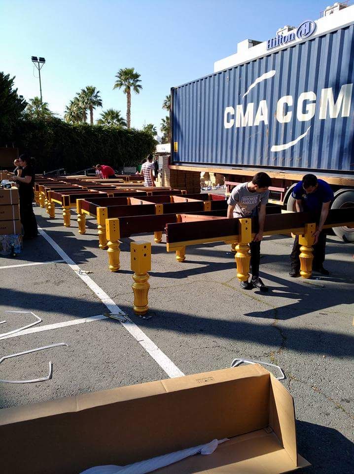 Mensen zetten houten constructies met gele steunen in elkaar naast een blauwe zeecontainer.