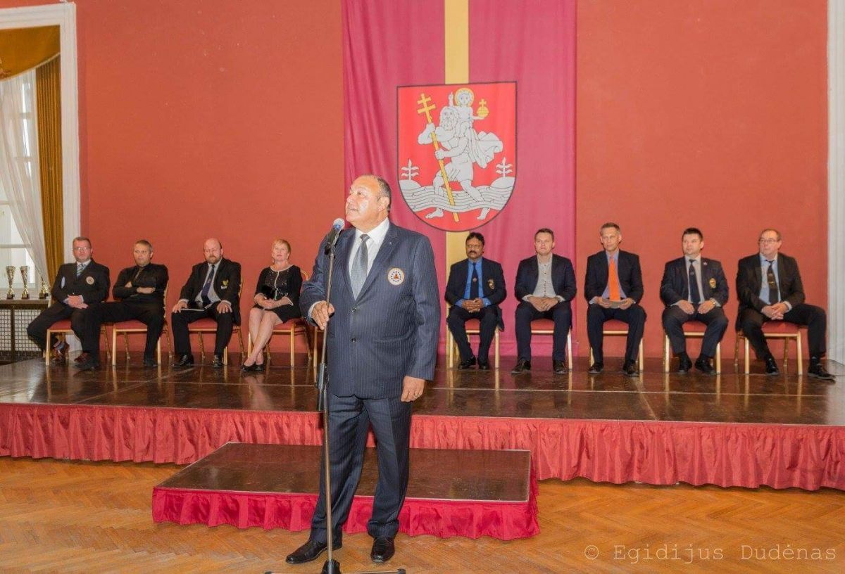 Een man spreekt tijdens een formeel evenement, met mensen in pak op een podium. Rode achtergrond, wapenschild.