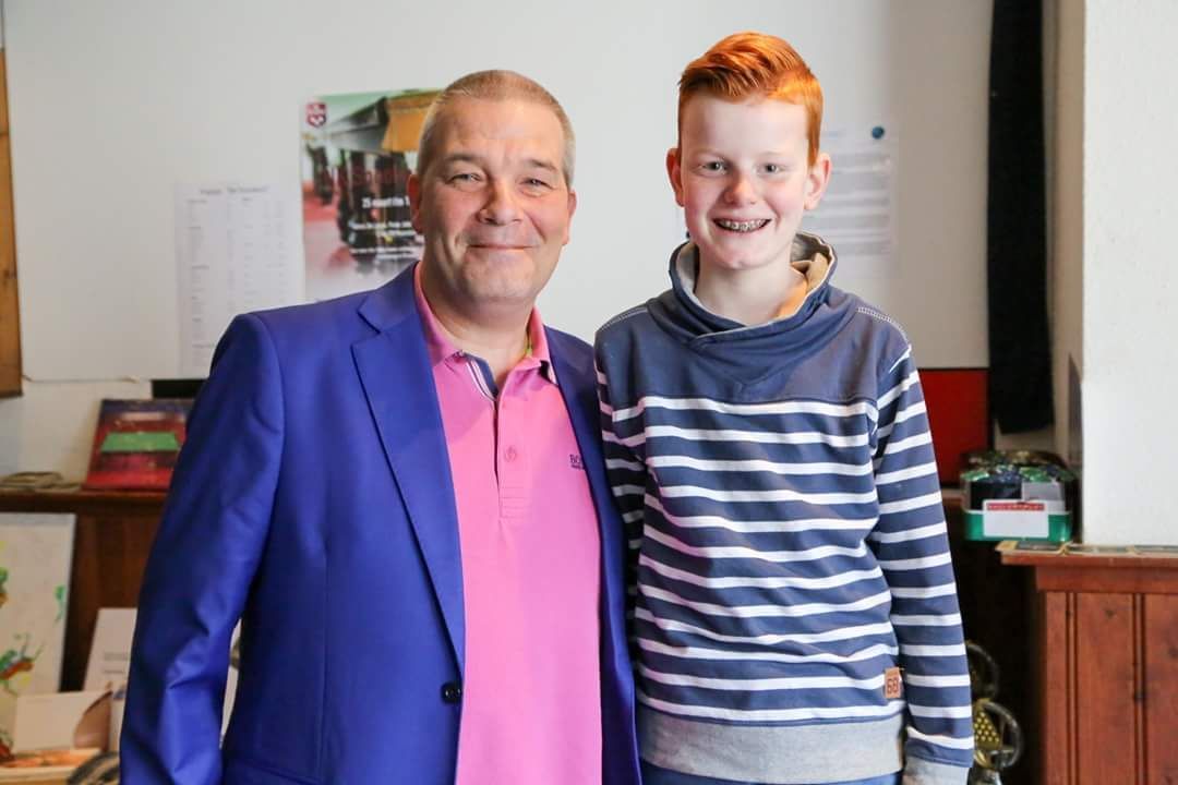 Man in blauw pak en roze shirt met lachende jongen met rood haar en bretels. Binnen, lachend.