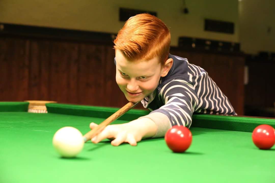 Jonge roodharige jongen speelt pool en mikt op een stoot. Groene tafel, rode ballen, binnen.