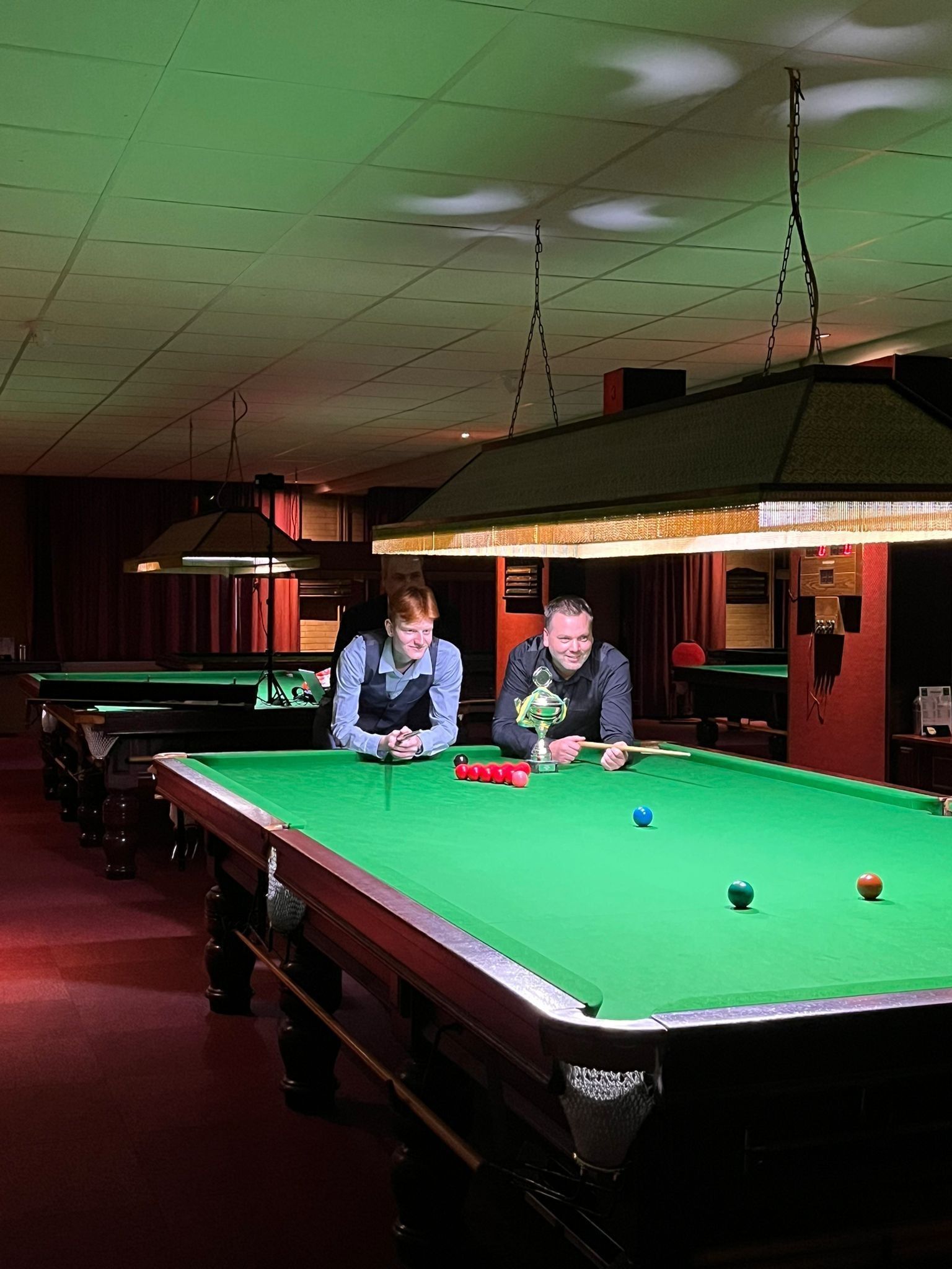 Twee mensen leunen over een snookertafel in een schemerige kamer, in een rood-groene kleurstelling.