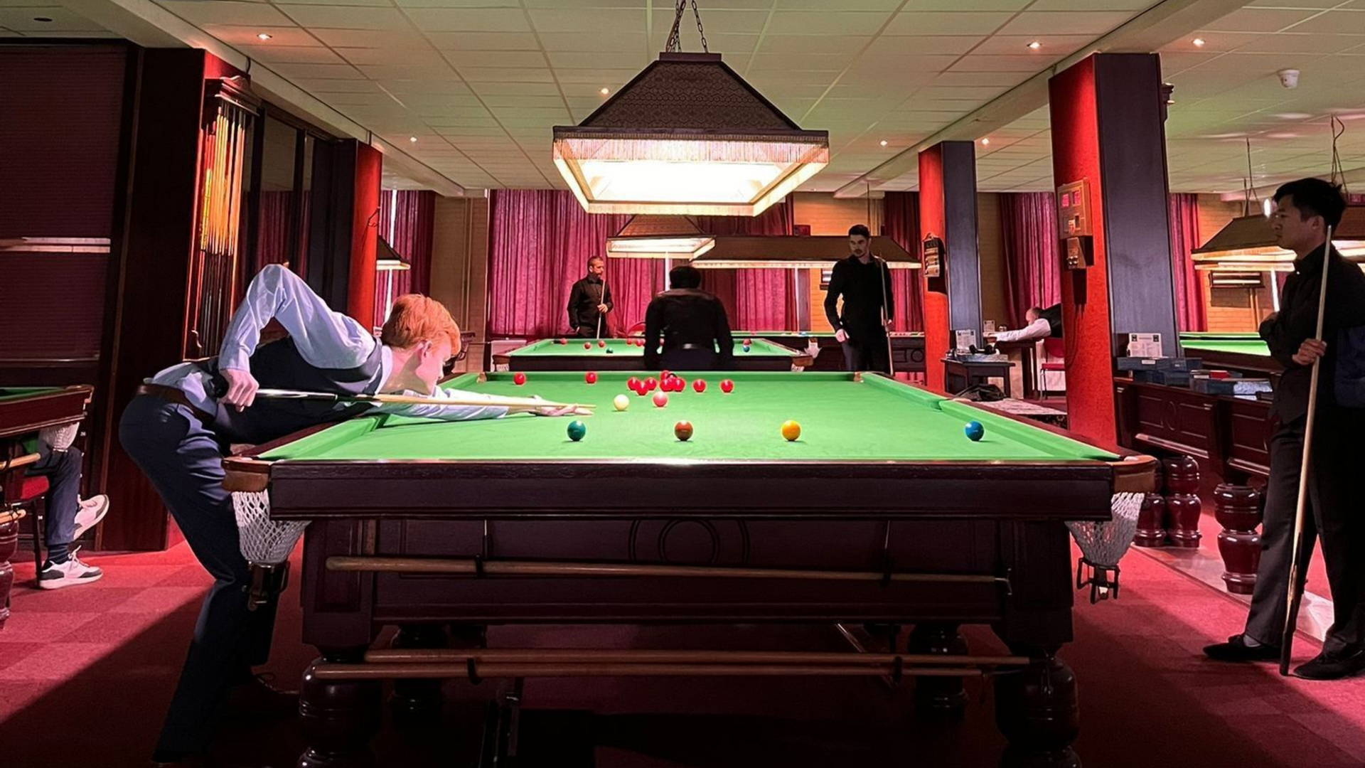 Man leunt over een snookertafel en richt op de ballen; hij staat met zijn clubs klaar terwijl anderen spelen.
