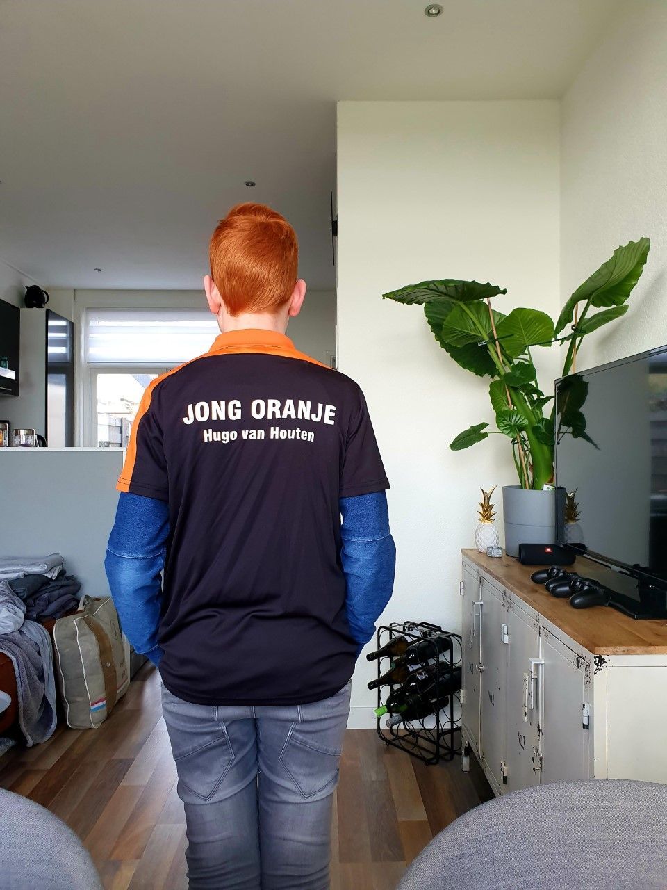 Jongen met rood haar, gekleed in een donkerblauw Jong Oranje-shirt, staat binnen met zijn gezicht van de andere kant af.