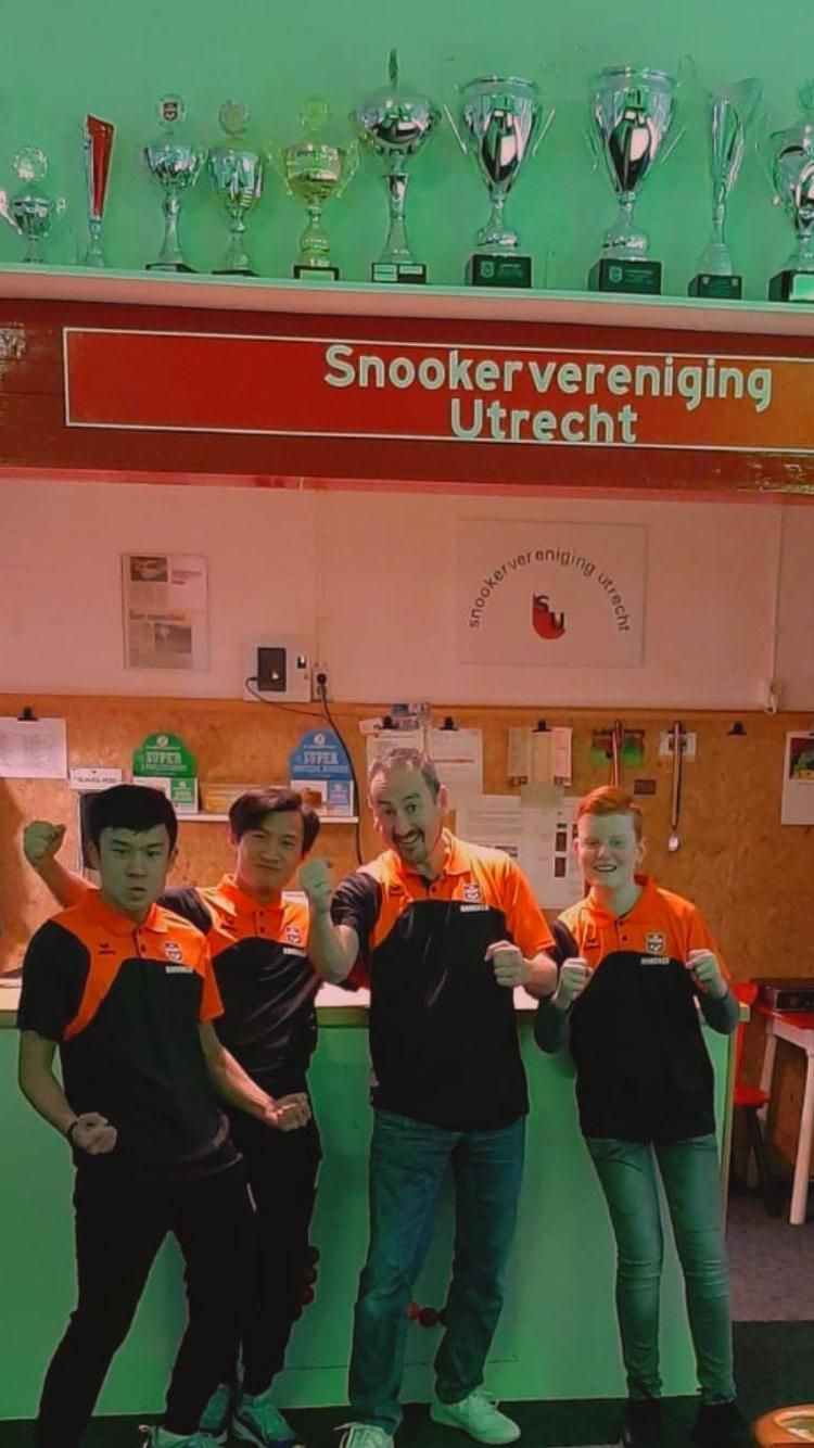 Vier mensen poseren enthousiast in een snookerclub, met trofeeën op een plank erboven; bord Snooker Vereniging Utrecht.