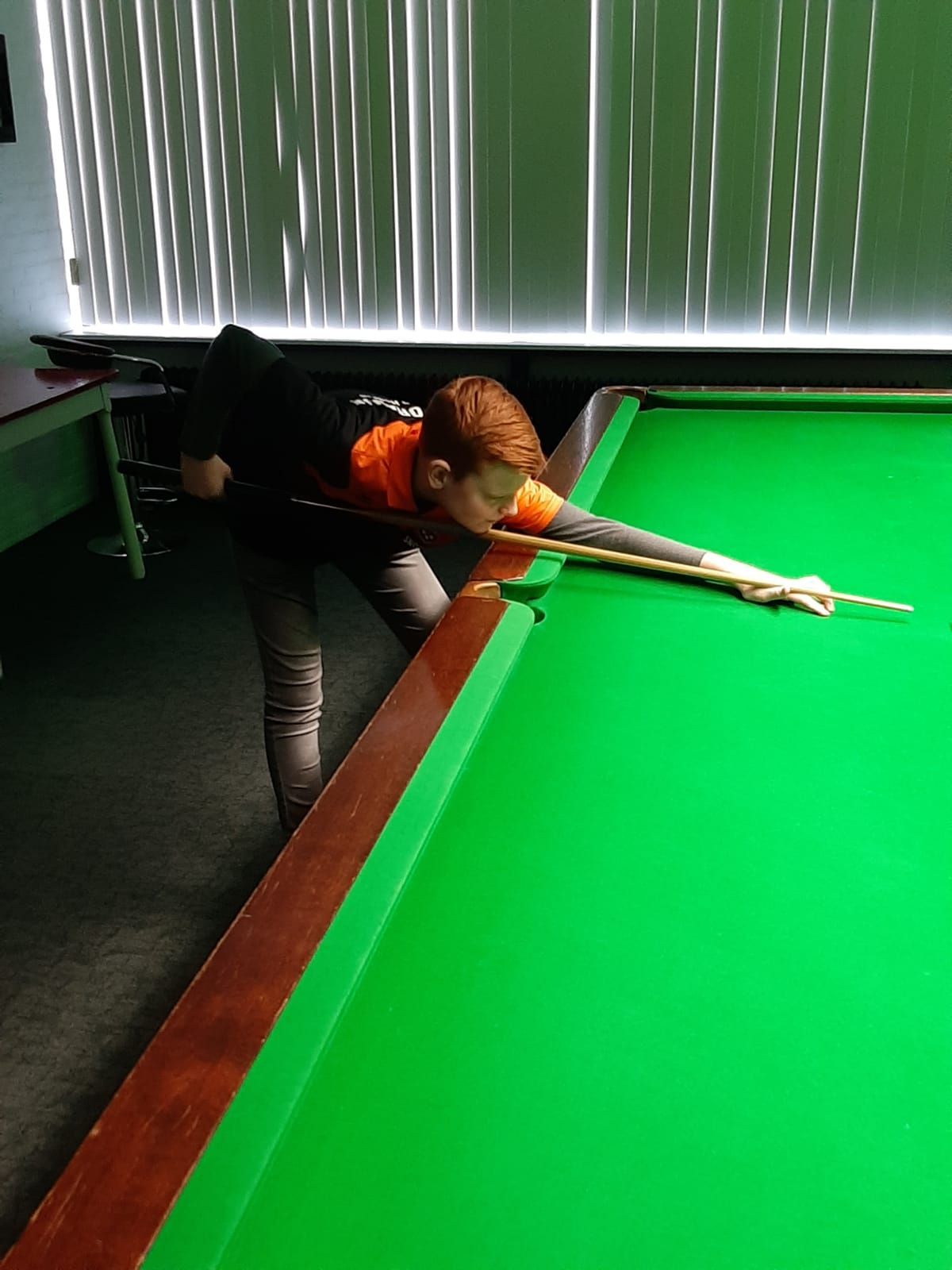 Een persoon met rood haar speelt snooker en richt de keu op een groene tafel in een slecht verlichte kamer.