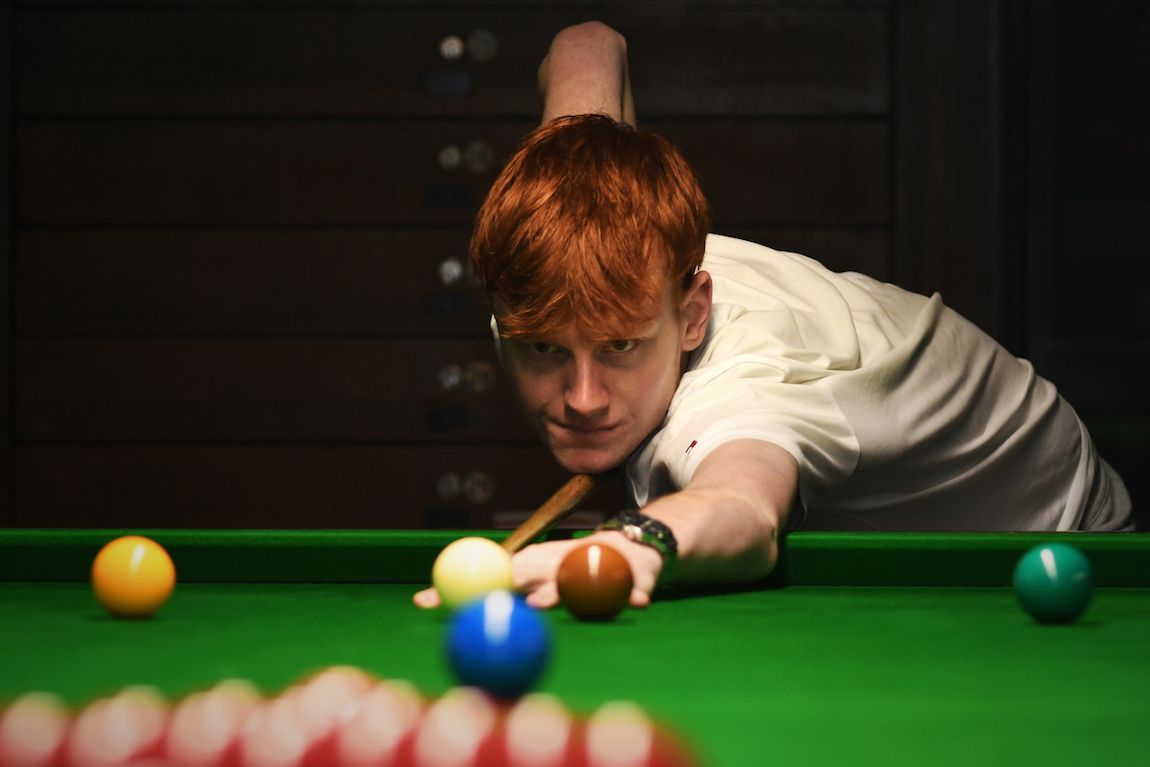 Snookerspeler met rood haar bereidt zich voor op een stoot, met zijn keu naast de speelbal, op een groene tafel.
