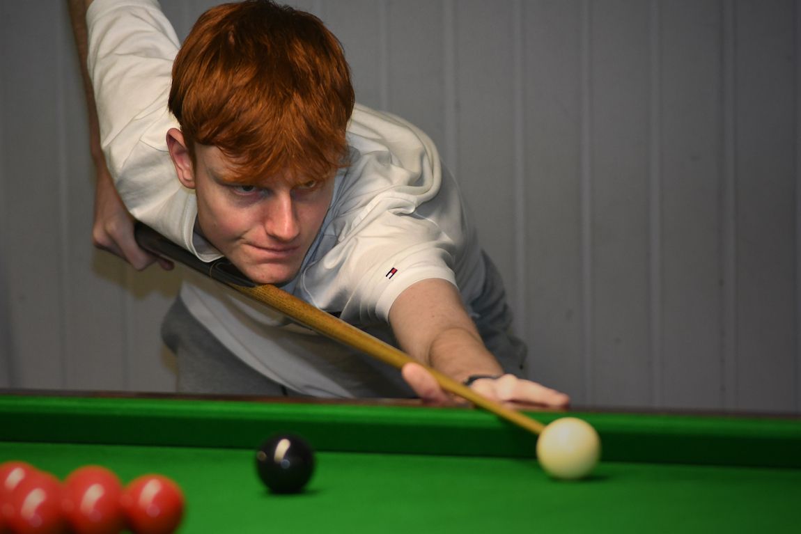 Een jongeman met rood haar richt een keu op een witte biljartbal op een groene tafel van vilt.