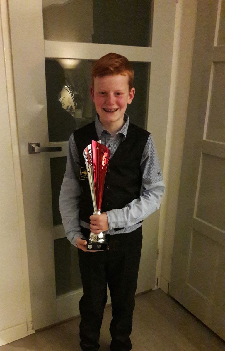 Een jongen met rood haar, gekleed in een zwart vest, houdt een trofee vast. Lachend staat hij voor een deur.