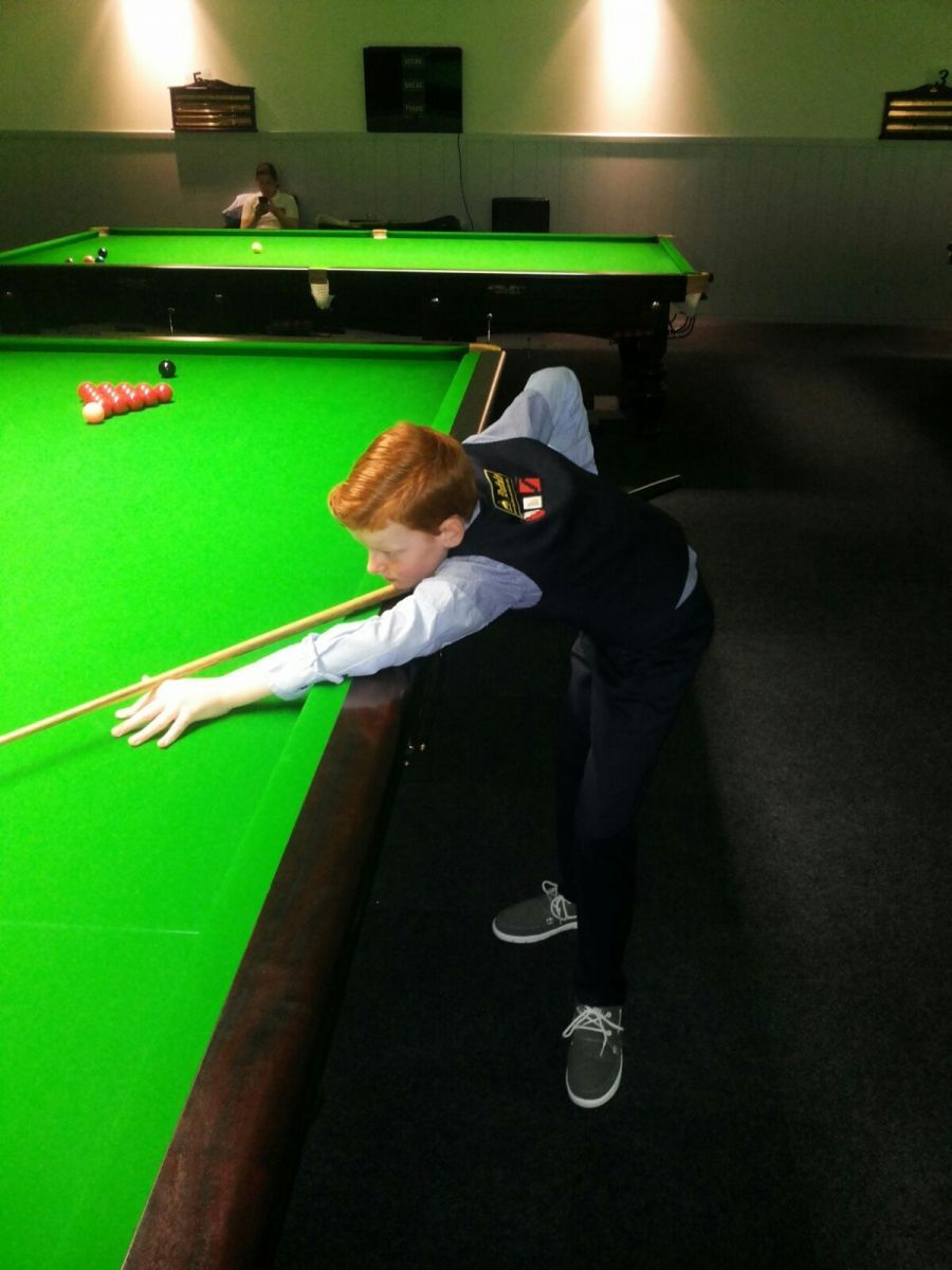 Een jonge roodharige in formele kleding richt een keu op een snookerbal op een groene tafel in een snookerhal.