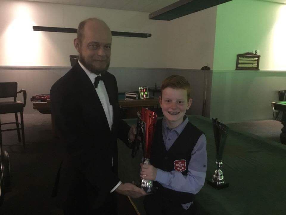 Man in smoking overhandigt trofee aan lachende jongen in vest aan een snookertafel.