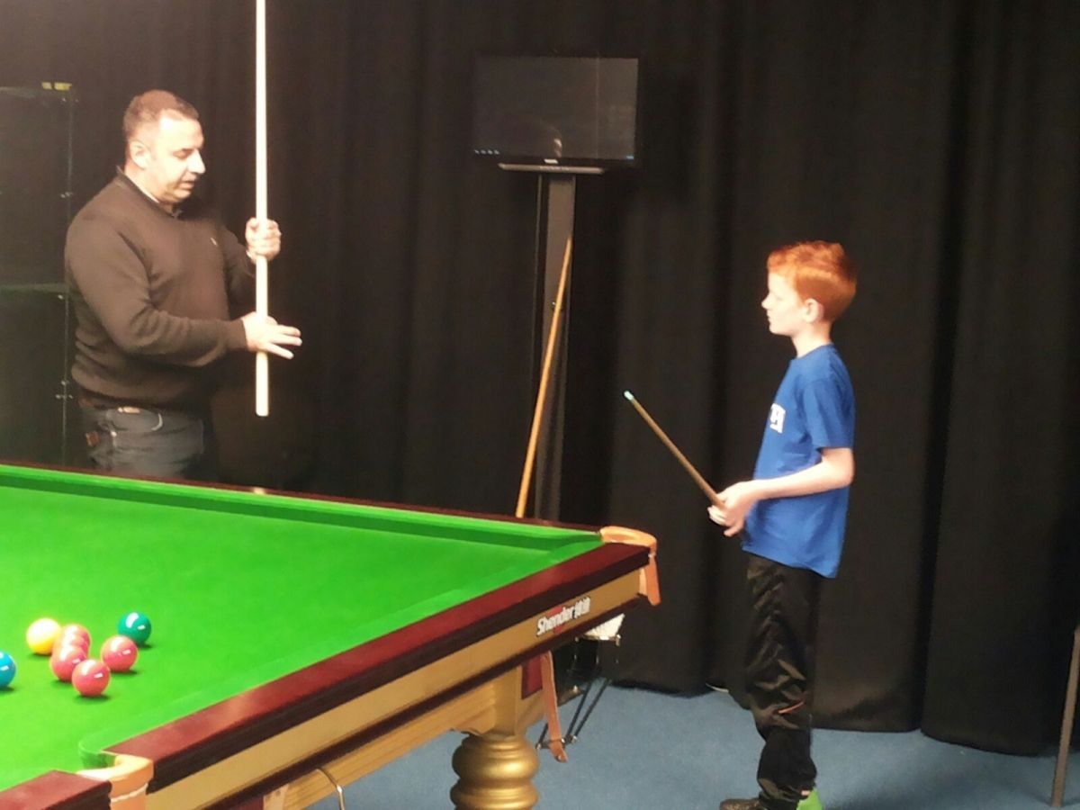 Man leert jongen snookeren. Man houdt keu vast; jongen richt op een andere. Groene tafel, zwarte achtergrond.