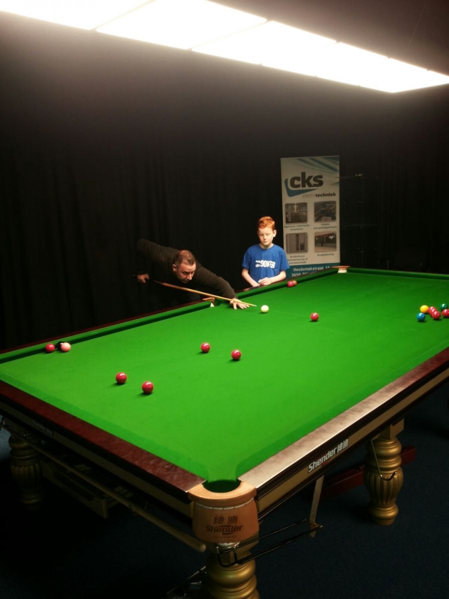 Een man bereidt een bal voor op een snookertafel, onder toeziend oog van een jongen in een blauw shirt. Donkere kamer, groene tafel.