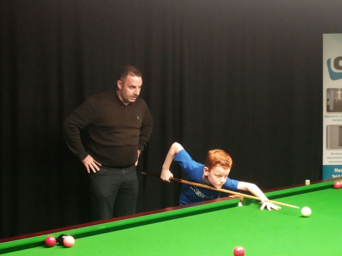 Een jongen in het blauw maakt een snookerbal klaar, terwijl een man toekijkt. Zwarte achtergrond, groene tafel.