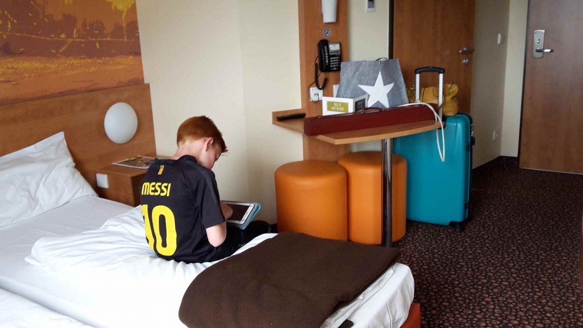 Jongen in Messi-shirt gebruikt tablet op hotelbed, bagage in de buurt.