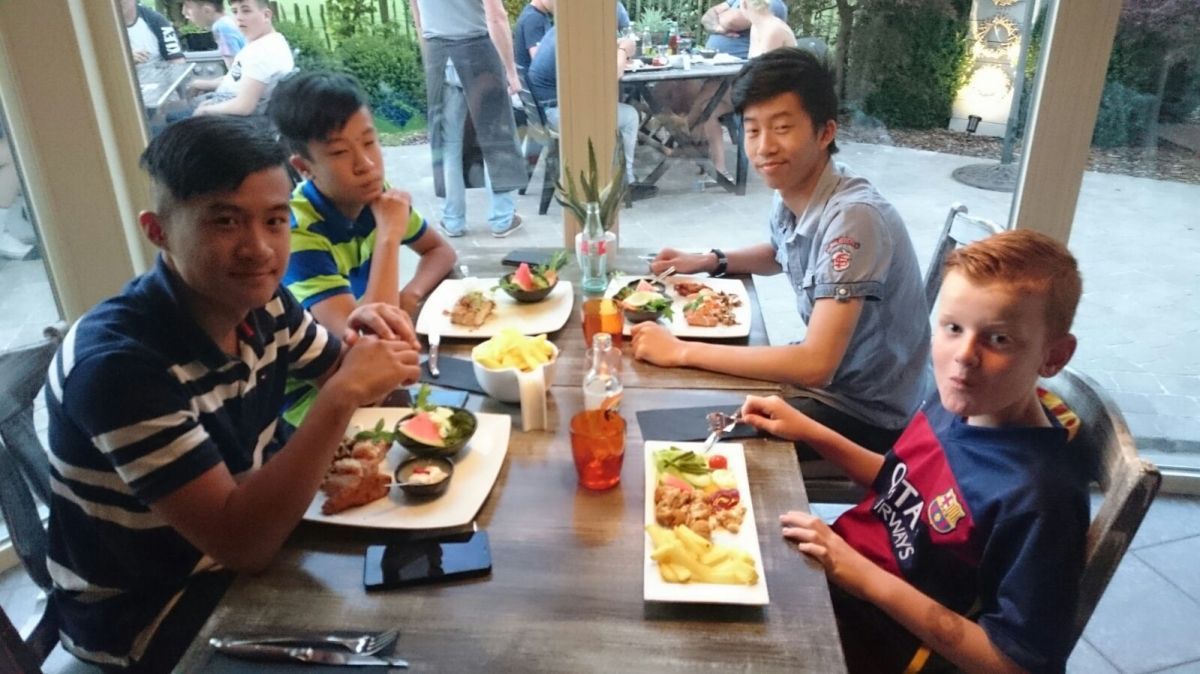 Vier jongens aan een tafel, etend. Eén jongen draagt ​​een voetbaltenue. Restaurantsetting.