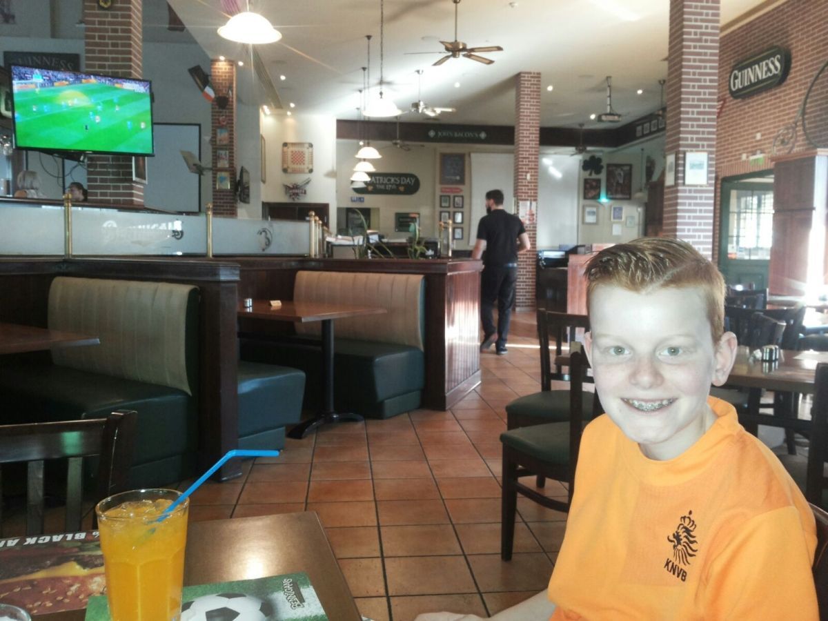 Jongen lacht naar de camera in een Ierse pub, drankje voor zich, tv op de achtergrond.