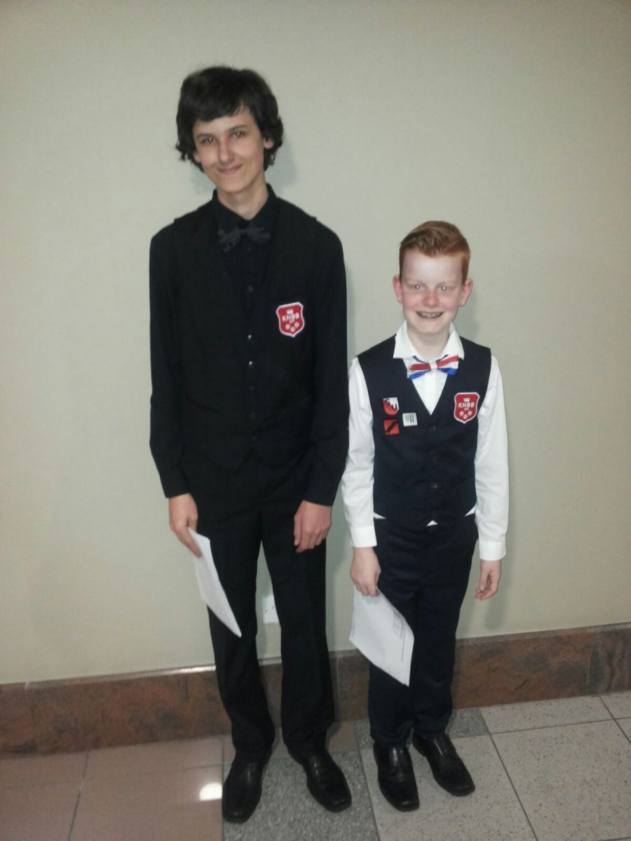 Twee jonge jongens in formele kleding, lachend en met papieren in hun handen, staan ​​binnen.