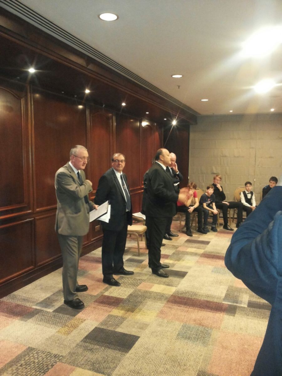 Vier mannen in pak staan ​​te praten; sommigen zitten op de achtergrond. Interieur.