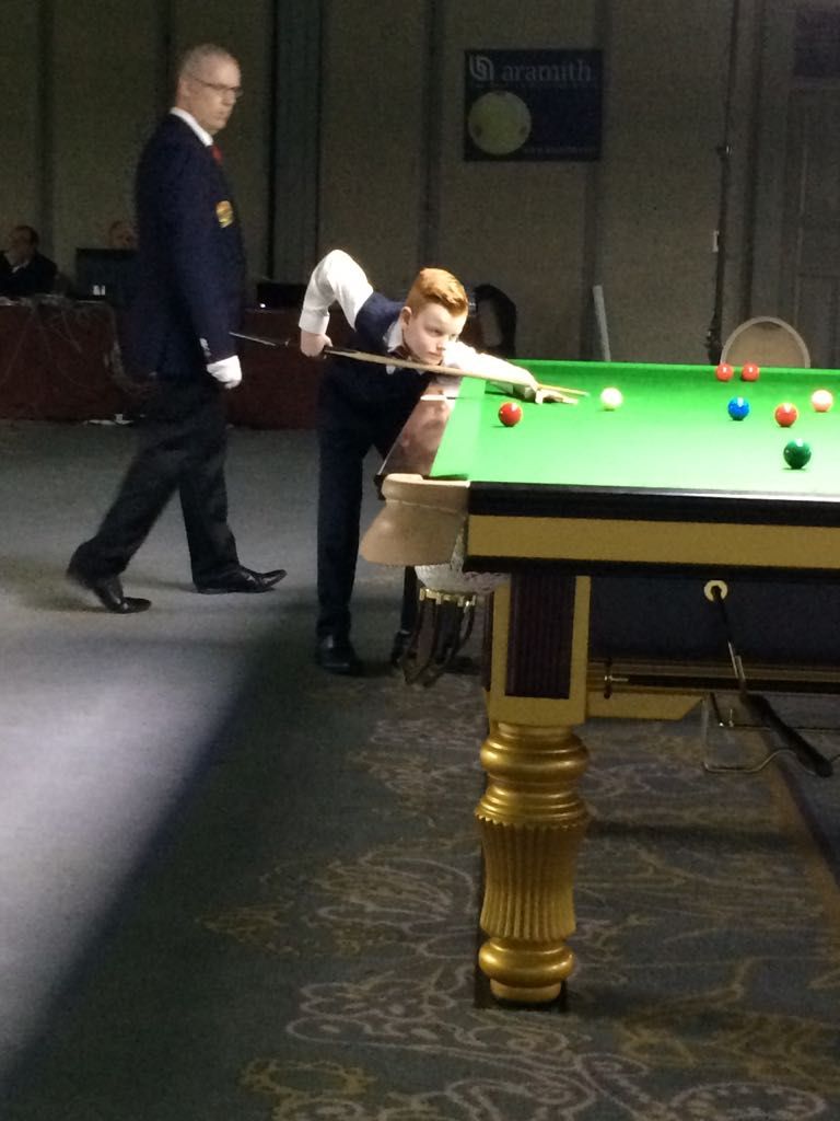Een jongere die aan het snookeren is, richt op de bal, terwijl de scheidsrechter toekijkt.