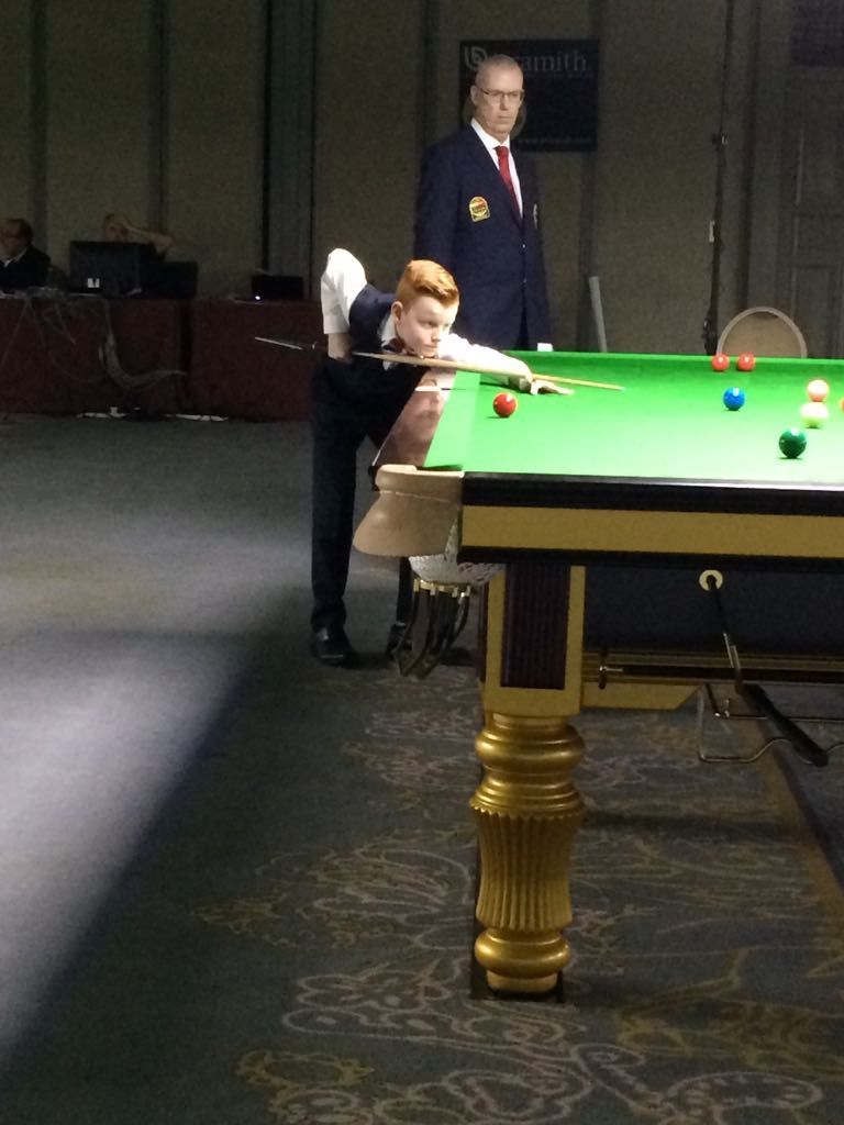 Snookerspeler maakt een slag klaar, keu uitgestoken. Scheidsrechter kijkt toe. Groene tafel, donker pak, indooropstelling.