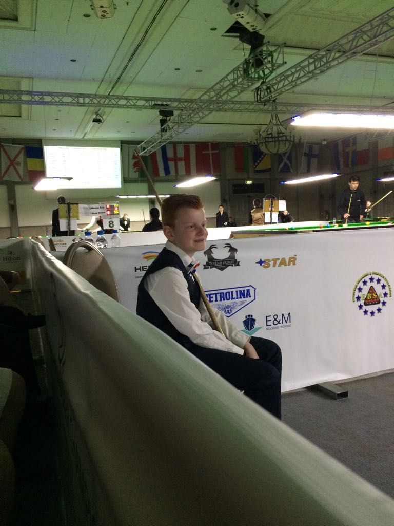 Een glimlachende jongeman in formele kleding zit aan een snookertafel. Binnenlocatie met vlaggen en logo's.