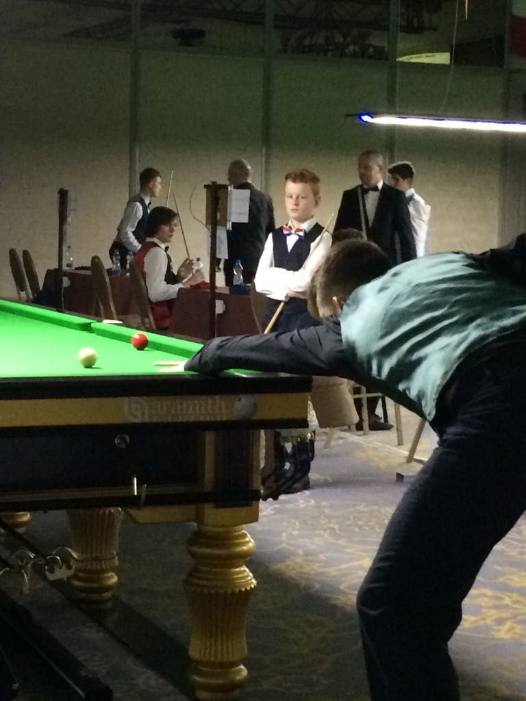 Een persoon die snooker speelt, concentreert zich op één slag; toeschouwers kijken toe in een donkere zaal.