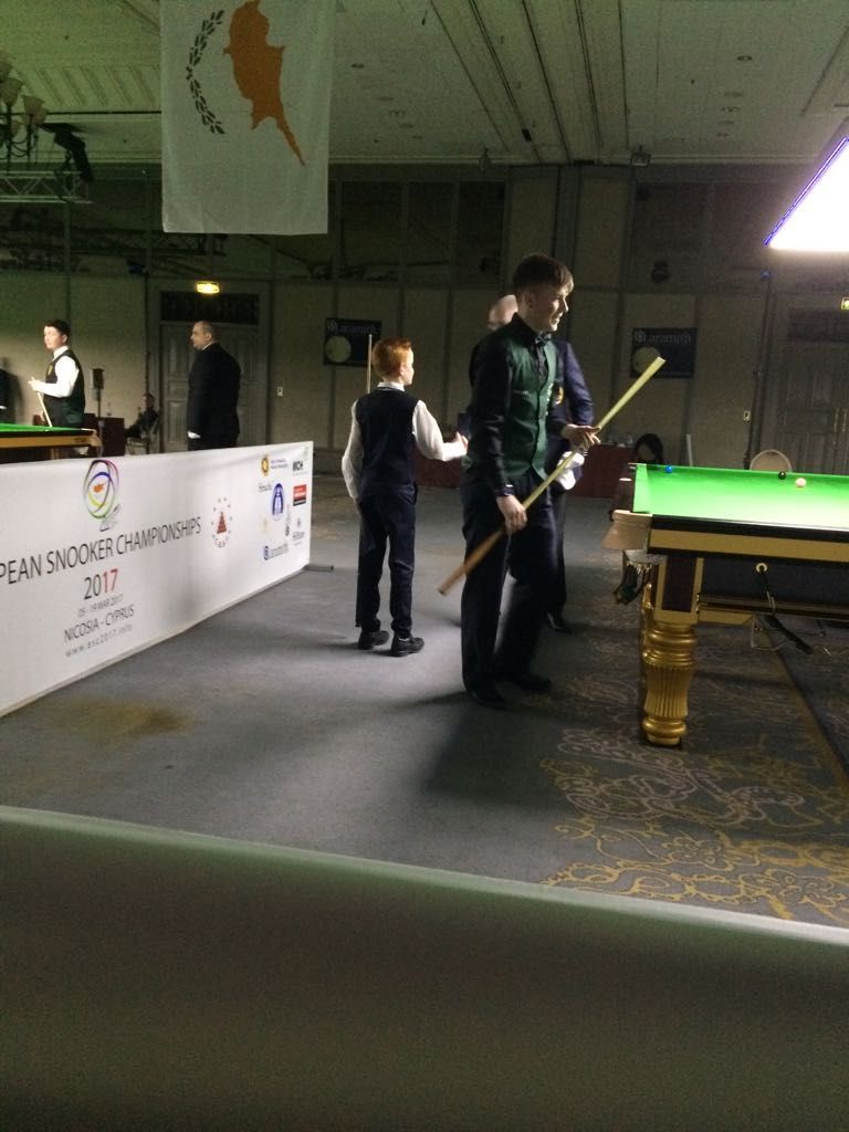 Snookerspelers in een toernooisetting. Een jonge jongen staat met een keu naast een volwassen speler. De vlag van Cyprus is zichtbaar.