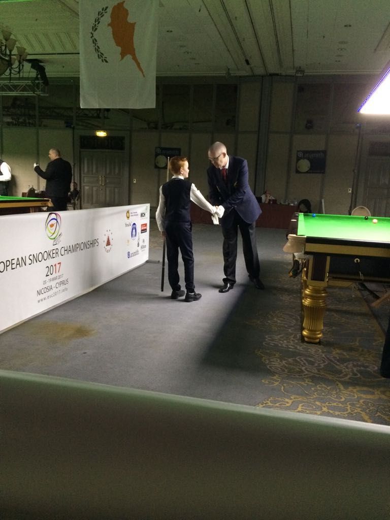 Twee snookerspelers schudden elkaar de hand voor een wedstrijd in een toernooihal; de vlag van Cyprus op de achtergrond.