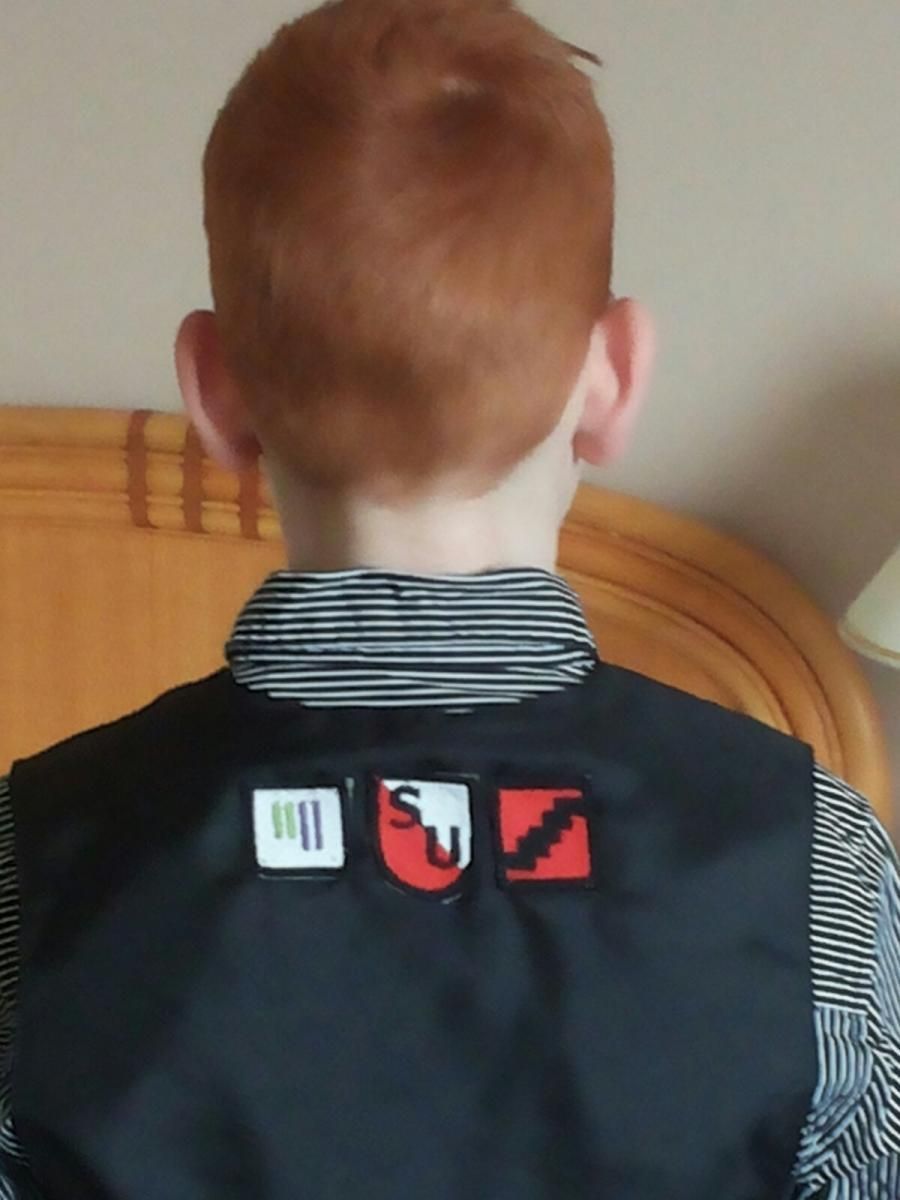Achterkant van een persoon met rood haar, die een zwart vest draagt ​​over een gestreept shirt met logo's op het vest.