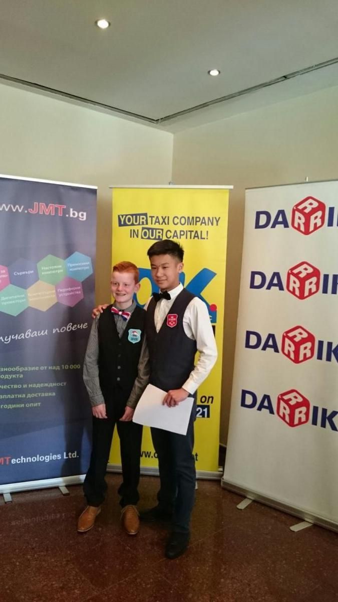 Twee jongemannen poseren voor een foto op een evenement met spandoeken. De een heeft rood haar en een vest, de ander houdt een papier vast en draagt ​​een vlinderdas.