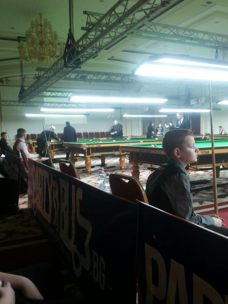 Jongen kijkt naar een snookerwedstrijd in een zaal met meerdere tafels, spelers en toeschouwers.