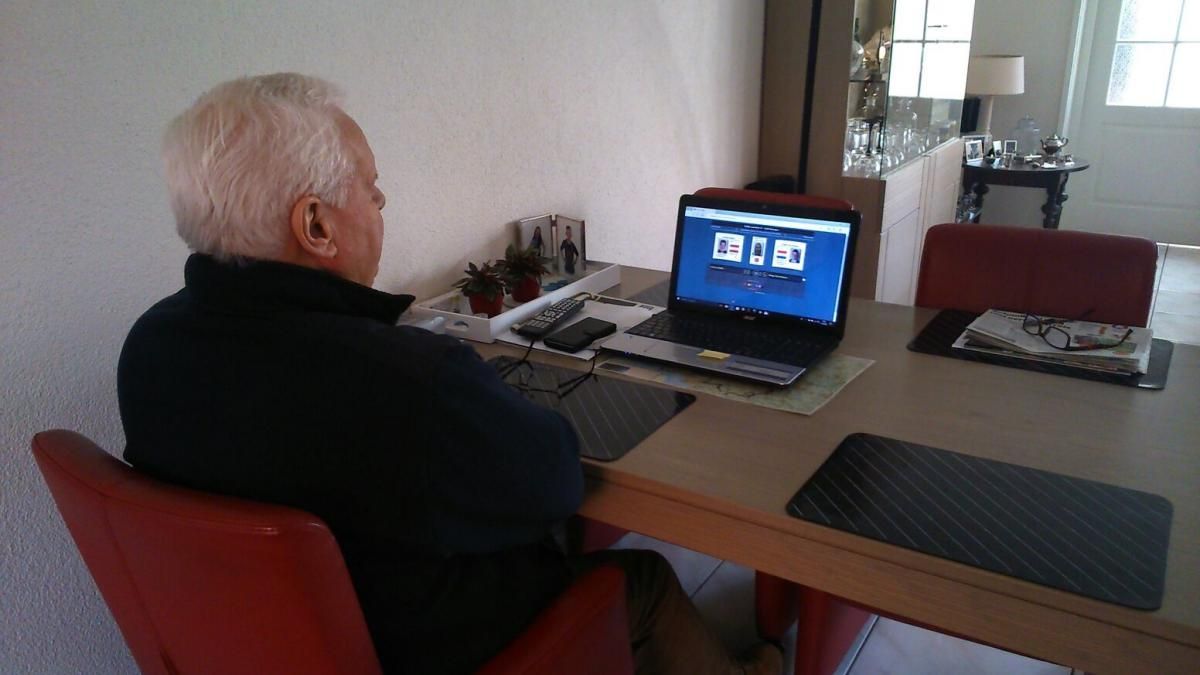 Een oudere vrouw met wit haar zit aan een tafel en kijkt naar een laptop. Ze bevindt zich in een kamer met een rode stoel.