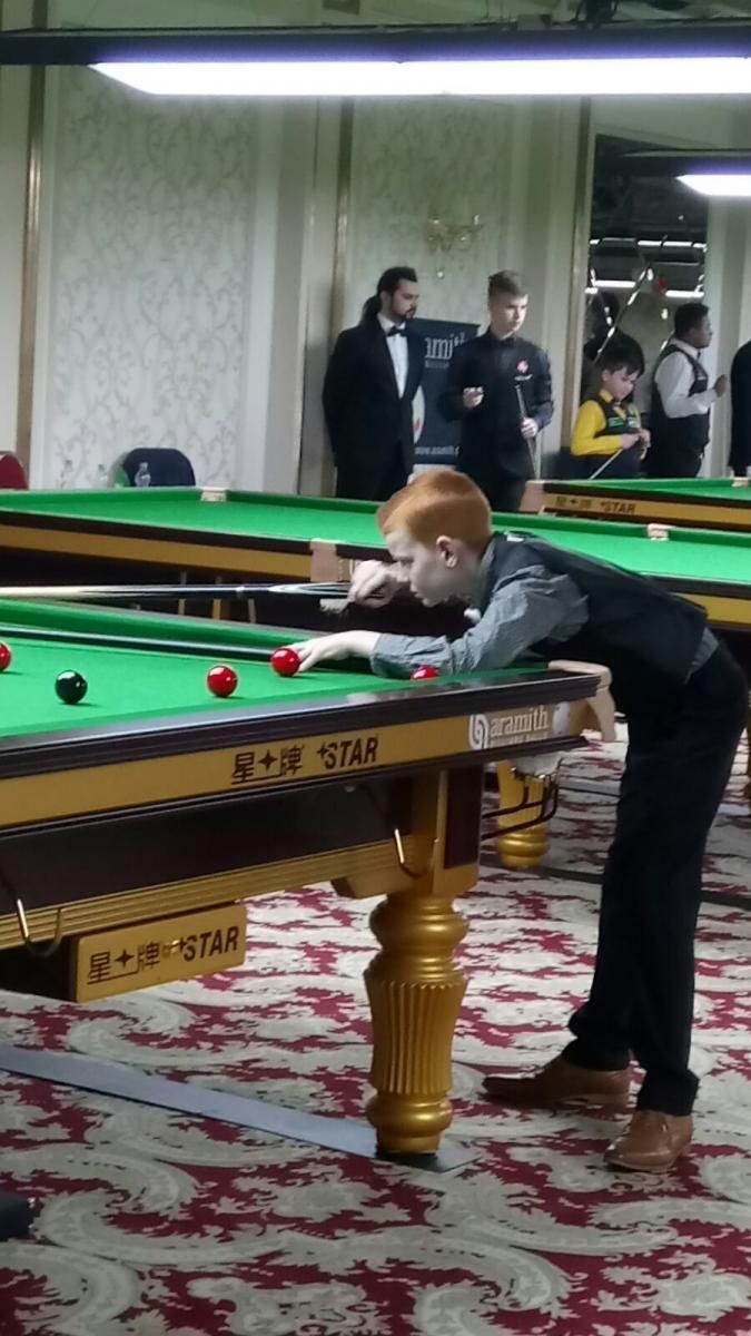 Een jonge roodharige in een vestje bereidt zich voor op een potje snooker aan een snookertafel in een zaal vol toeschouwers.
